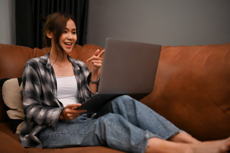 Attractive young Asian female is having a great conversation with her friends during video calling through her laptop computer in her living room.の写真素材