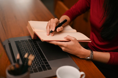 A professional businesswoman is listing something on her notebook while working at her office desk. cropped and close-up imageの写真素材
