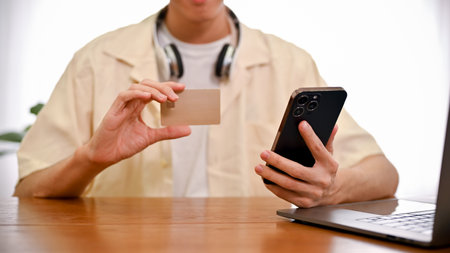 A young Asian man sits at his desk, using smartphone and holding a credit card. Online payment,...