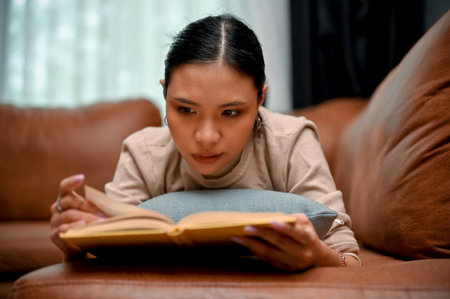 Attractive and gorgeous young Asian female concentrating on reading a book while relaxing, lying on...