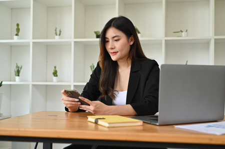 Attractive and beautiful millennial Asian businesswoman or female boss sits at her office desk using her smartphone to send emails or text.の写真素材