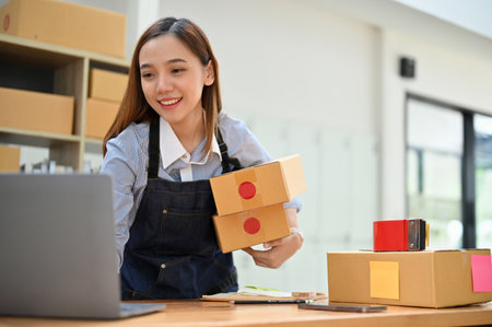 Successful millennial Asian female online shop owner or small business startup checking customer's online orders on laptop while preparing shipping packages. e-commerce business conceptの写真素材