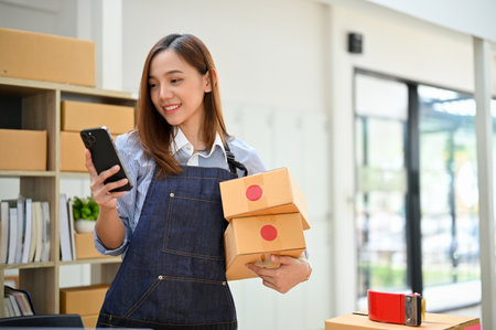 Gorgeous young Asian female online shop owner or business startup using her smartphone while holding her shipping packages in her stock office. E-commerce business startup conceptの写真素材
