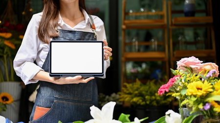 Beautiful young Asian female florist or floral shop owner in apron showing or holding a digital tablet white screen mockup, standing in front of her flower shop. cropped shotの写真素材