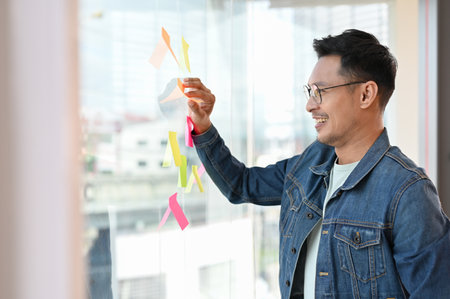Smart adult Asian businessman or male marketing worker looking at a memo notes or sticky notes on...