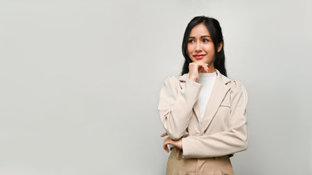 Confident millennial Asian female or businesswoman in casual suit thinking, planning, hand on chin, standing isolated over grey background. copy space for your advertising contentの写真素材