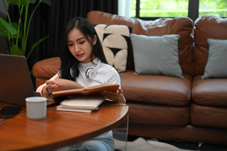 Smart young Asian female college student concentrating doing homework, reading a book, researching information in textbook, sitting in her living room.の写真素材