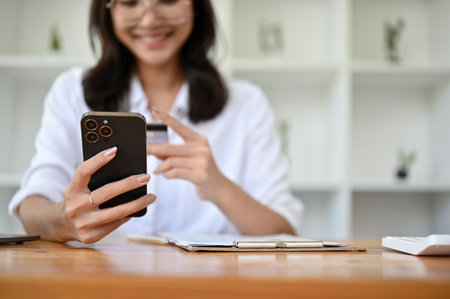 Attractive millennial Asian female sits at her desk, holding a smartphone and a credit card. close-up image. Online payment, credit card payment, mobile banking, online shopping, cashless society.の写真素材