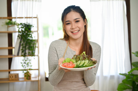 Attractive and smiling young asian female wearing apron, holding a plate of fresh organic green salad. Healthy food and lifestyle conceptの写真素材