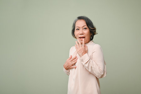 Surprised and excited 60s senior Asian woman in casual standing against isolated green studio background. shocked pose for advertising your big shopping deal.の写真素材