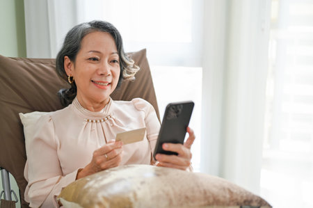 Beautiful Asian aged-woman using an online mobile banking application to pay her bills while relaxes sitting on her comfy armchair in her living room.の写真素材