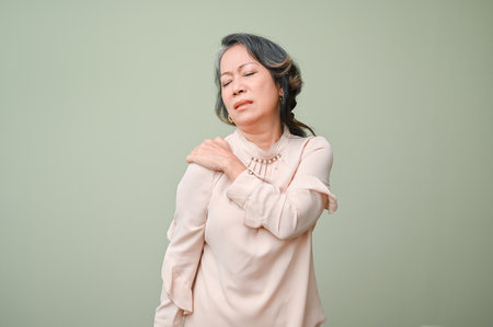 60s aged Asian woman suffering from her shoulder pain, massaging her shoulder, standing over isolated green studio background.の写真素材