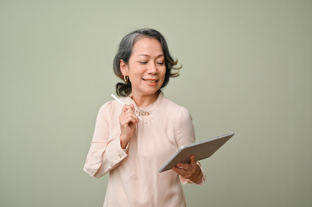 Happy and cheerful aged-asian woman in casual clothes holding a digital tablet and stylus pen, standing isolated over green background.の写真素材