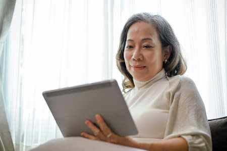 A beautiful Asian-aged woman watches a video clip online on a digital tablet touchpad in her comfortable living room.の写真素材