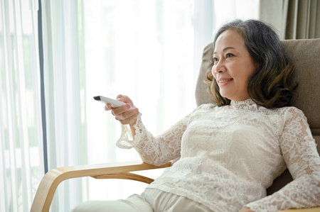 An attractive Asian middle-aged woman enjoying watching a game show on TV in her cozy armchair in the living room.の写真素材