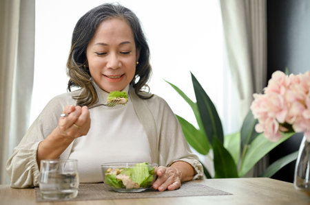 Beautiful and healthy Asian-aged woman eating healthy green salad with chicken breast in her dining...