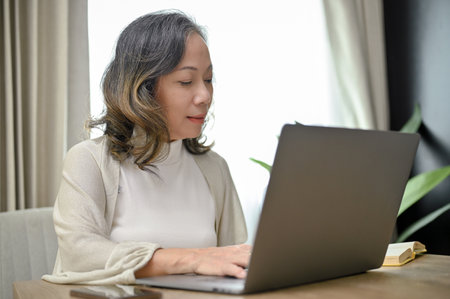 Attractive and professional Asian-aged woman or female entrepreneur working from home, using laptop computer in her living room.の写真素材