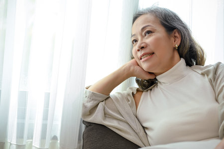 Attractive beautiful Asian-aged woman relaxes in her comfortable living room, sits on sofa looking out the window, daydreaming about healthy life and happiness.の写真素材