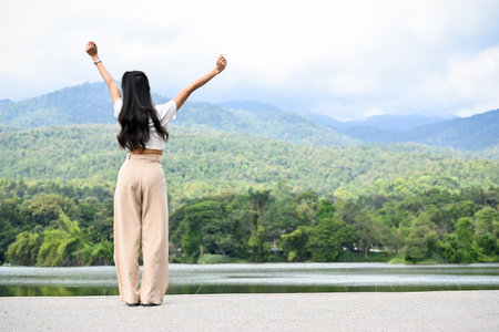 Young Asian female raising hands with freedom and happiness expression, breathing fresh nature air, standing by the lake with green mountain view. back viewの写真素材
