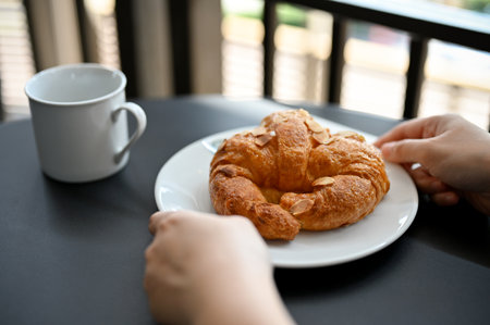 A female hands holding a delicious fresh French croissant with almond slice on a white plate serve with hot coffee or cocoa.の写真素材