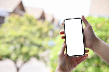 A female's hands holding a smartphone white screen mockup over blurred outdoor view with trees in...