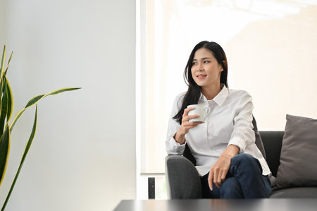 Attractive young Asian woman relaxes sipping morning coffee while chilling in her living room.の写真素材