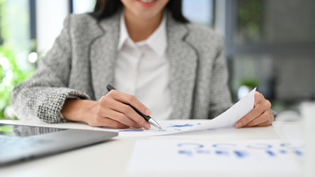 Beautiful millennial Asian businesswoman or female financial analyst checking financial data on the report, analyzing business financial data, working in the office. cropped imageの写真素材