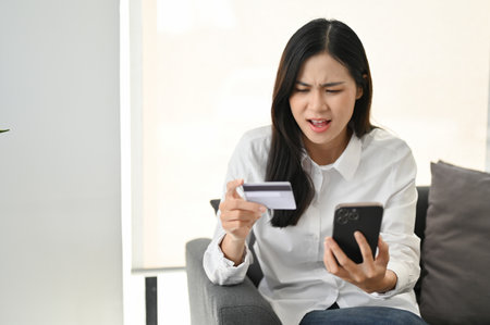 Dissatisfied Asian woman sitting on sofa, looking at her smartphone screen, having problem with...