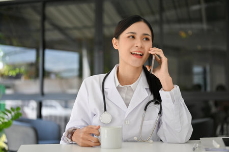 Attractive and professional young Asian female doctor in white gown talking on the phone with someone at her office desk.の写真素材