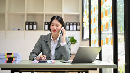 Beautiful Asian businesswoman talking on the phone with her client while working at her office desk.の写真素材