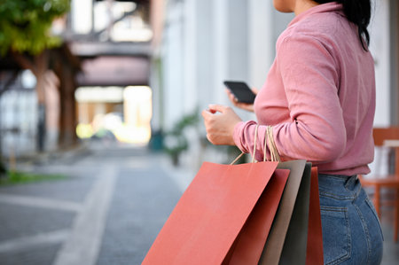 Beautiful young Asian woman in casual clothes carrying her shopping bags and using her smartphone...