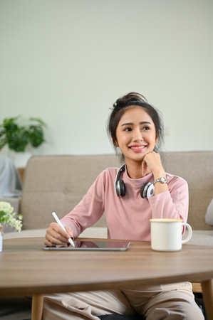 Portrait, Beautiful young Asian female using her digital tablet at a coffee table in her living room, hand on chin, smiling, daydreaming and looking out window.の写真素材