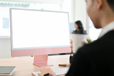 Young Asian male office worker working at his desk, looking at the computer screen, typing on keyboard computer. PC desktop computer white screen mockup. back viewの写真素材