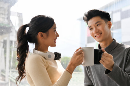 Lovely young Asian office-couple standing by the window, having afternoon coffee break together.