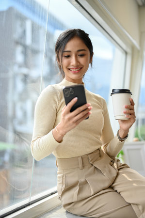 Portrait, Attractive and charming young Asian female in casual clothes sitting on window bench, enjoying her coffee and using her smartphone.の写真素材