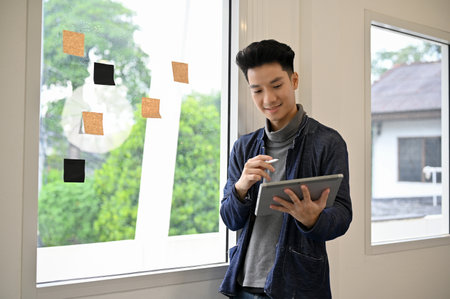 Smart and creative young Asian male marketing employee in casual clothes stands by the window...