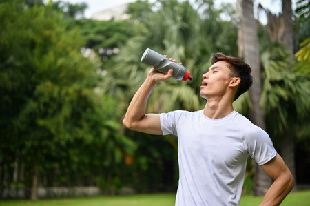 Handsome, thirsty and sweaty millennial Asian athletic man in sportswear drinking water or protein drink from bottle after training in the park.の写真素材
