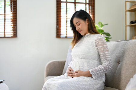 Beautiful and happy millennial Asian pregnant woman relaxes sitting on sofa, touching her belly. pregnancy, motherhoodの写真素材