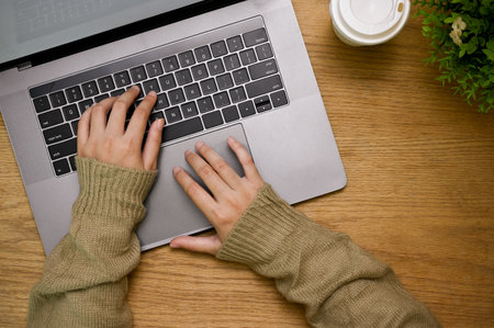 A female in cozy sweater typing on notebook keyboard at her wooden desk, laptop computer, coffee cup...