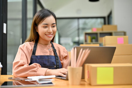 Beautiful young Asian female e-commerce business startup checking her orders on laptop computer in the stock room.の写真素材