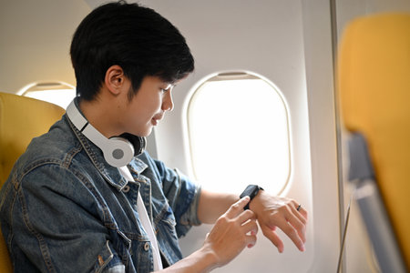 Handsome young Asian male passenger sits at a window seat in economy class, checking a certain time...