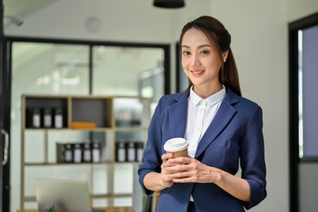 Confident and elegant young Asian businesswoman or female CEO in stylish business suit holding a cup of coffee, standing in her modern office.の写真素材