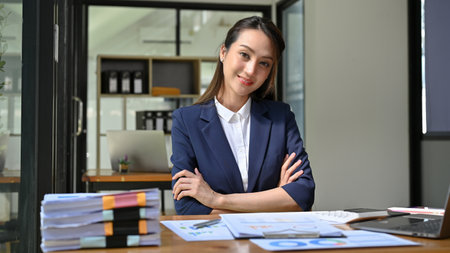 Attractive millennial Asian businesswoman or female manager sitting at her desk, arms crosses, smiling and looking at camera.の写真素材