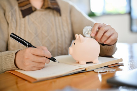 A female making her revenue and expenses accounts on a book and insert coin into a piggybank.の写真素材