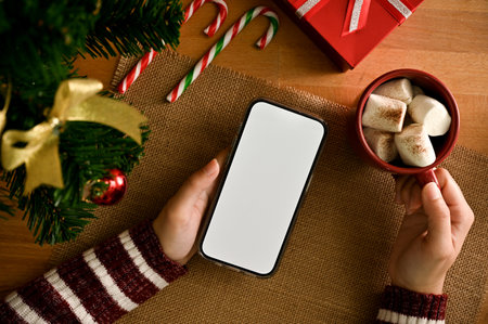 A female hands holding a smartphone white screen mockup and a hot chocolate with marshmallows on...