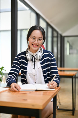 Portrait, Beautiful and charming young Asian female with headphones doing homework, writing something on her notebook while sitting in the cafe co-working space.の写真素材