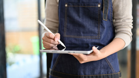 A female waitress wearing an apron takes a customer's order on her portable tablet. coffee shop worker, cashier, cafe owner. cropped and close-up imageの写真素材