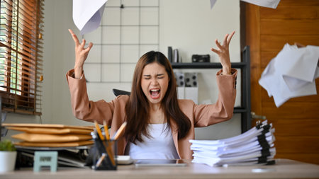 Angered, furious, crazy and mad millennial Asian businesswoman or female office worker screaming, shouting and throwing up papers at her office desk. Depression, overworked, failure.の写真素材
