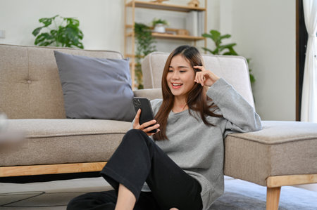 Relaxed and chilling young Asian female sits in her comfortable living room and uses her smartphone message with her friends or scrolls on her social media.の写真素材