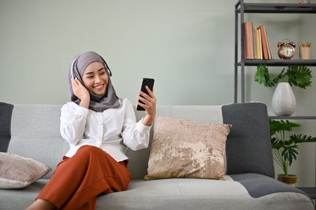 Attractive millennial Asian Muslim woman wearing hijab and headphones using her mobile phone, listening to her favorite song while relaxing in her living room.の写真素材